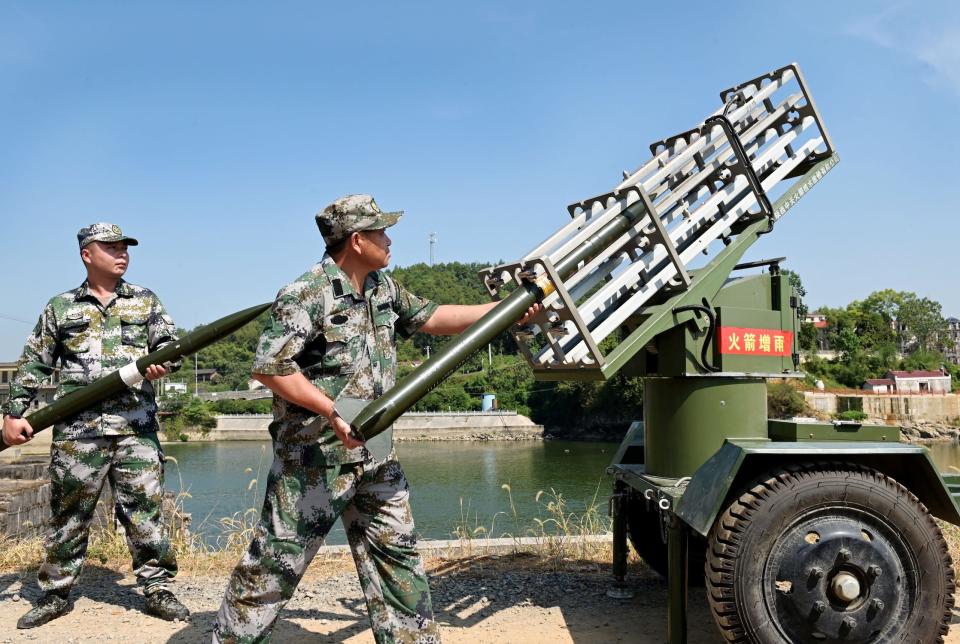 two men in camouflage fatigues loading long rocket-shaped tubes into a metal rack aimed at the sky atop a green platform on wheels