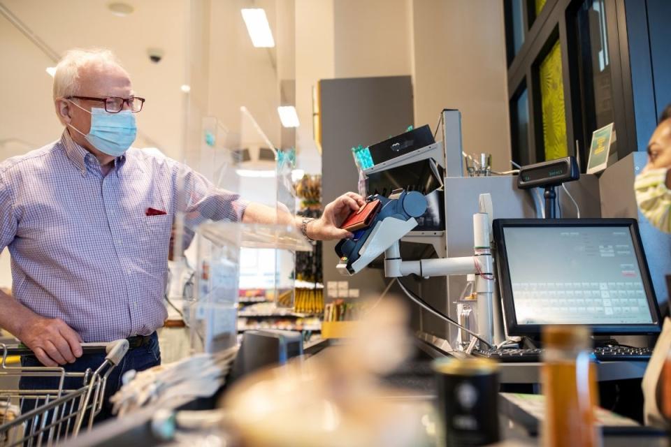 Senior shopper wearing face mask pays using phone at checkout.