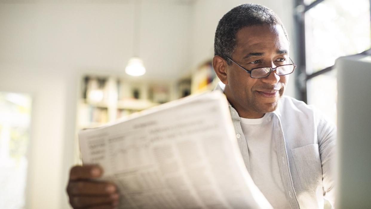 Senior man with newspaper using laptop.