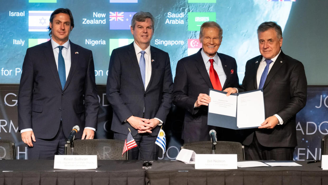  Four men in dark suits stand in front of a table with microphones and small flags on it. 