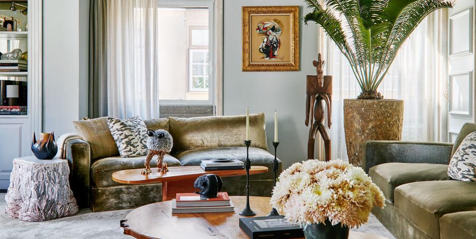 a living room with two sofas in moss green velvet with decorative pillows, a small and larger cocktail table with curvy edges, a terra cotta side table, female fertility sculpture, tall palm plant, light gray carpet