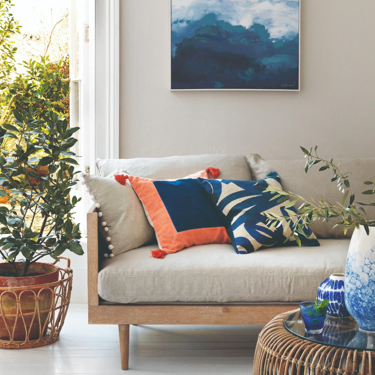  A living room with a sofa with a wooden frame, an abstract artwork hanging on the back wall and a potted tree placed next to the sofa. 