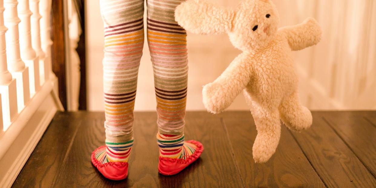 little girl wearing colorful striped leggings and moccasins and holding a soft cuddly stuffed bunny toy at the top of stairs at home