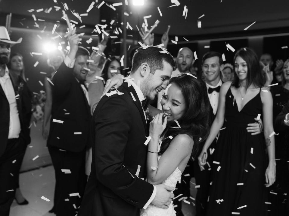 A bride and groom embrace as confetti explodes around them in black and white.