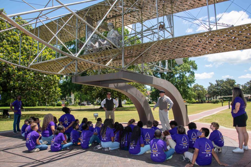 Elementary students from Bear Exploration Center For Mathematics, Science and Technology School perch in the shade of the Wright Flyer replica as they learn about the history of aviation from “Orville” and “Wilbur Wright,” May 13, 2022.