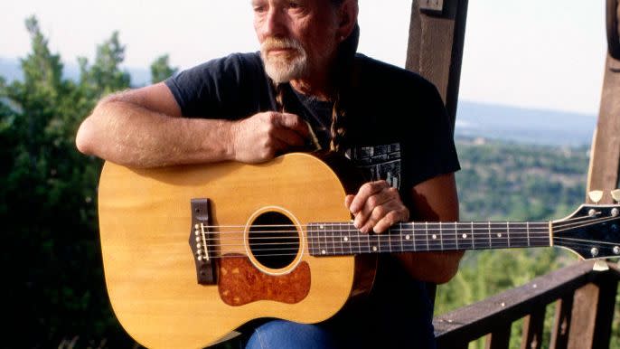 willie nelson standing against a fence and holding a guitar