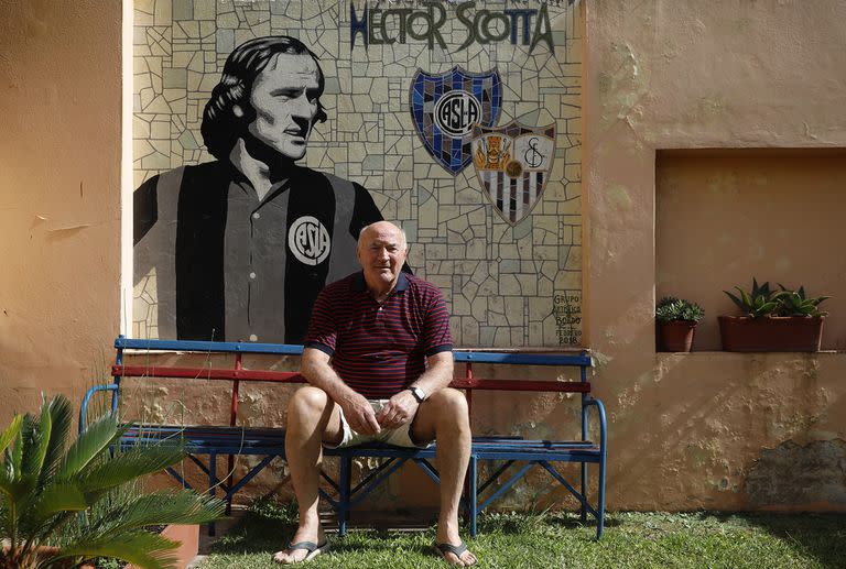 Héctor Scotta, en el jardín de su casa, sentado en un banco del Viejo Gasómetro y delante de un mural que lo homenajea, con los escudos de San Lorenzo y Sevilla, sus dos amores