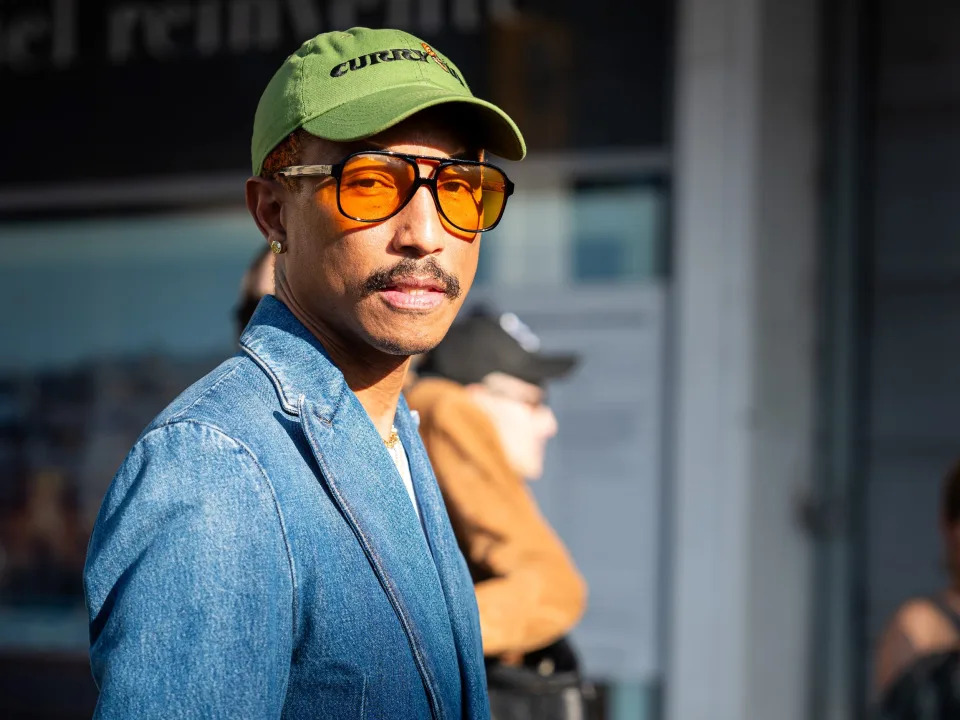 A man in a green cap, denim jacket and jeans, and a black, wide-frame sunglasses with orange lenses