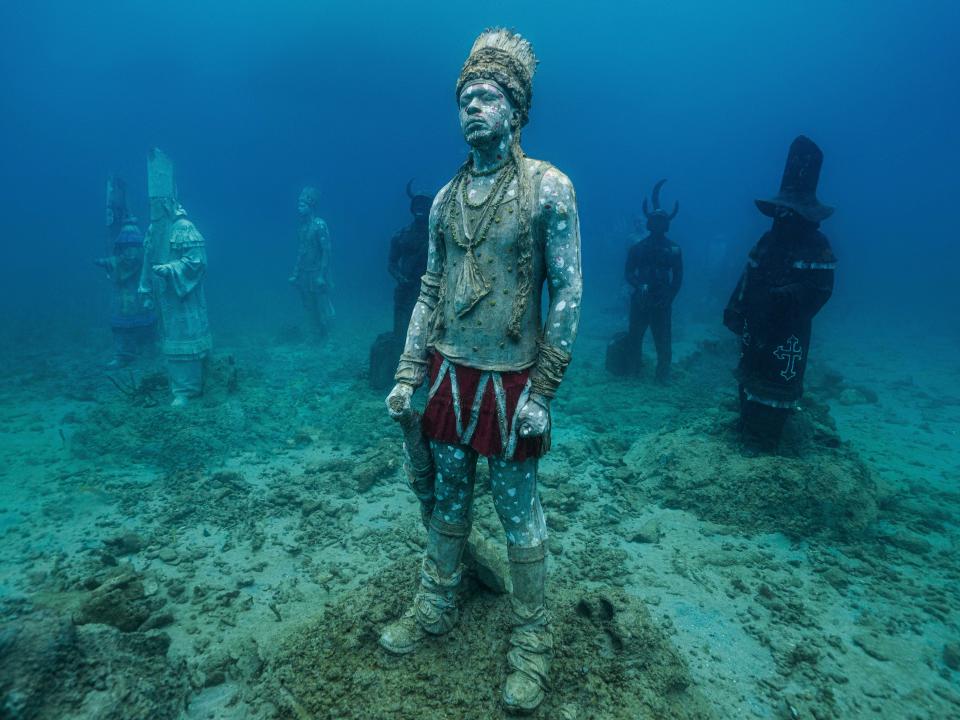 An underwater sculpture of a man with body and face paint wearing a headdress. Behind him are 6 other sculptures.
