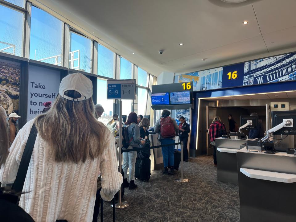 A line of people wait to board an American Airlines flight at New York's LaGuardia airport. In the foreground, a woman wearing a baseball cap and white sweater stands looking towards the gate, while other passengers stand holding suitcases and bags in the background.