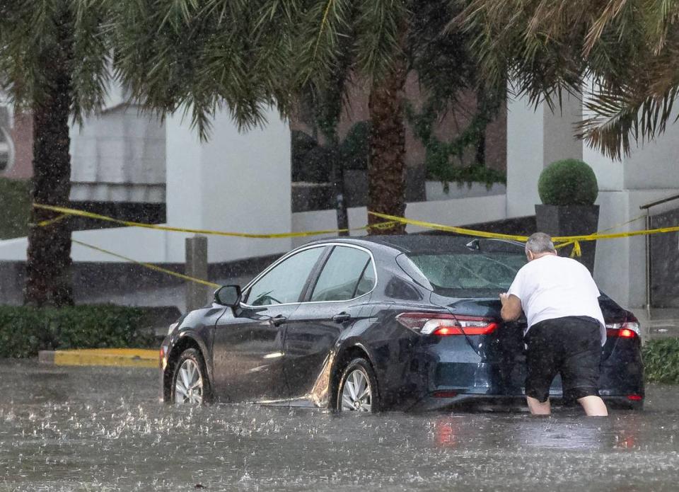 A person pushes a car through a flooded portion of Northeast 11th Street in downtown Miami as lightning and heavy rain falls over the area on Monday, June 19, 2023.