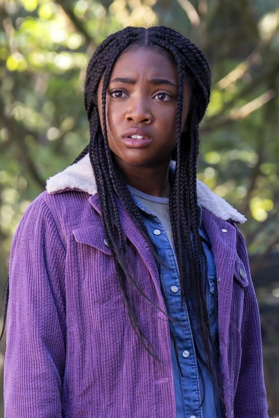 Leah as Annabeth in a layered outfit stands with a knife, looking serious, in a wooded area