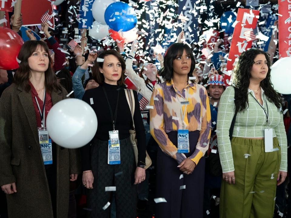four women, melissa benoist, carla gugino, christina elmore, natasha behnam, standing next to each other looking unamused. they're wearing DNC press credentials, and there are confetti and balloons falling around them