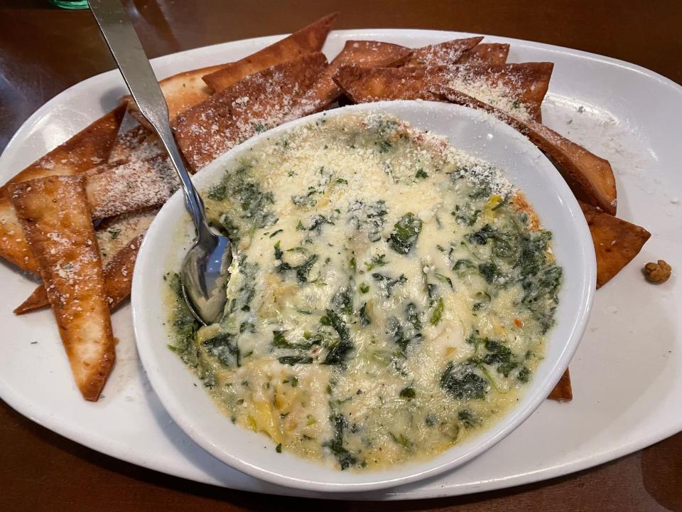 Bowl of spinach-artichoke dip with a spoon in it. Pieces of bread dusted with cheese sit beside the bowl on a platter