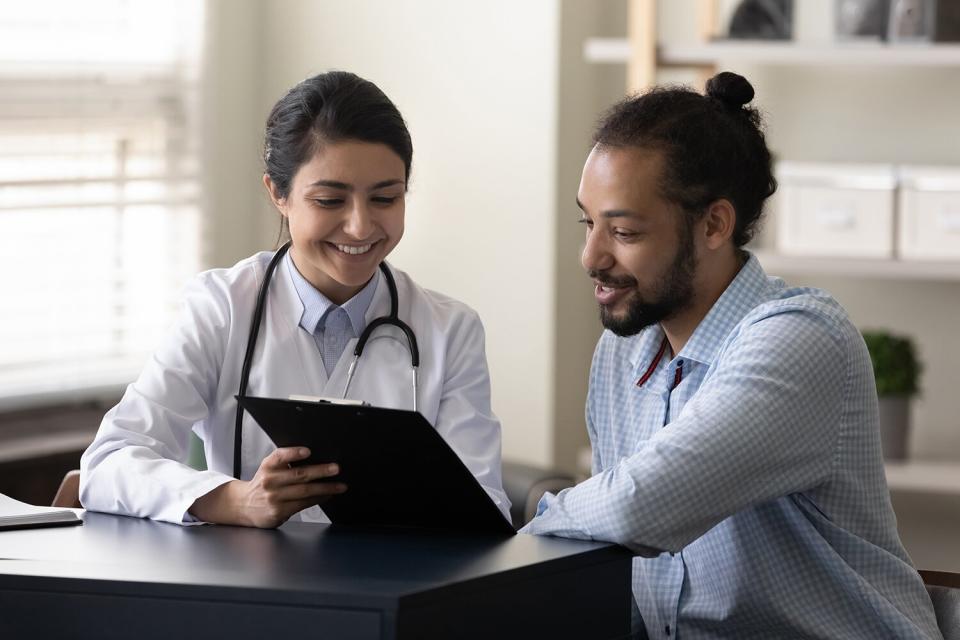 Happy young indian female gp doctor showing test result on clipboard to interested snake person african ethnicity male patient, discussing healthcare medical insurance or illness treatment at clinic.