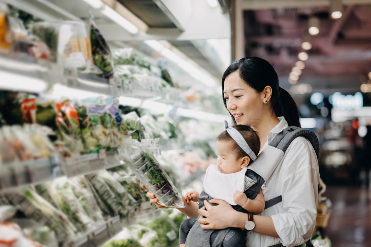 Asian mother carrying cute baby girl shopping for vegetables and grocery at the supermarket, illustrating a story on the best credit card for grocery shopping in Singapore.