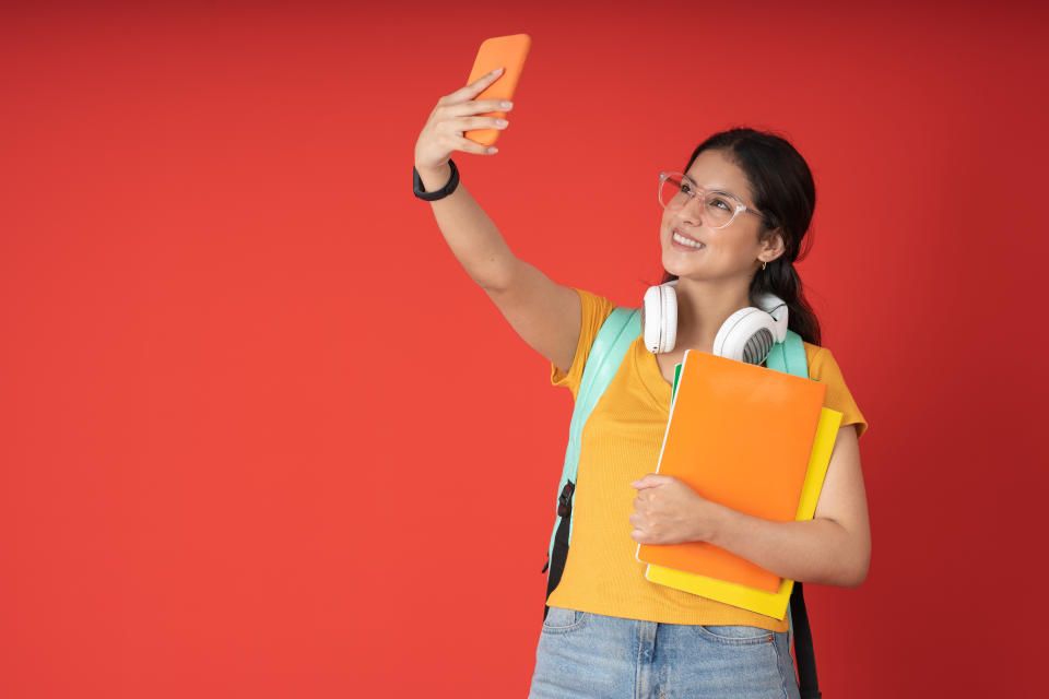 Young woman with glasses, taking a selfie smiling, while carrying her notebooks, on a red background.