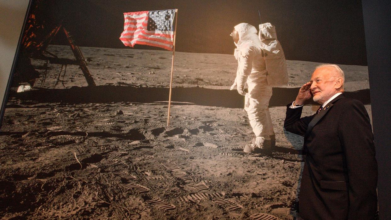  buzz aldrin salutes next to a poster-sized picture of himself. the poster shows buzz aldrin in a spacesuit saluting next to the american flag on the moon. at the left of the picture is the apollo 11 lunar lander. the surface of the moon is covered in bootprints 