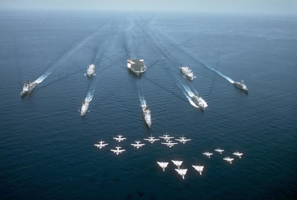 The USS America sails with its battle group while 16 aircraft fly overhead.