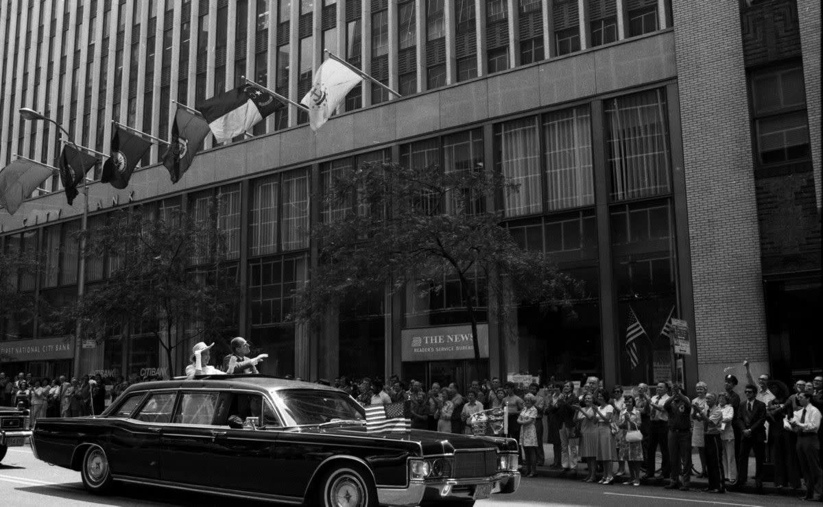 Flags of the 13 colonies flutter from The Daily News building as Queen Elizabeth II and Prince Philip wave to bystanders in New York City, circa 1976.