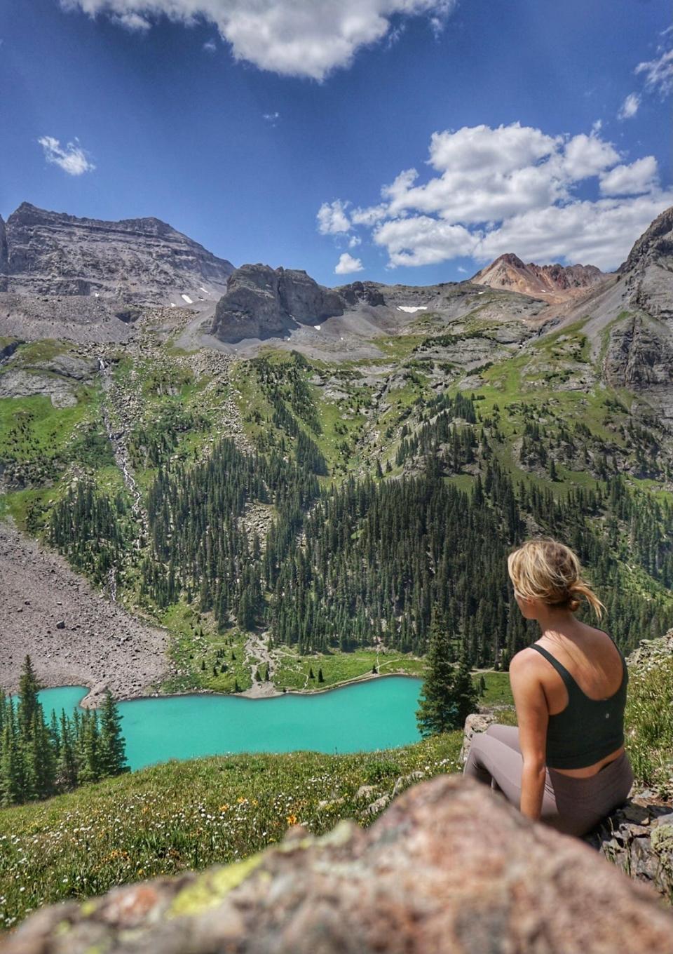A woman with her back to the camera facing a blue lake, mountains, and trees.