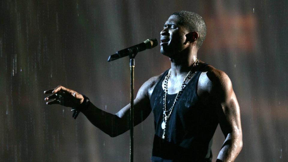 usher sings into a microphone on a stand while reaching one hand in front of him, he wears a black tank top, jeans, two necklaces, and a watch, water rains down on him