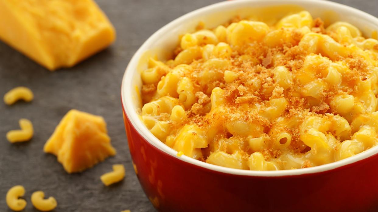 "Homestyle baked macroni and cheese in a red bowl with shredded cheese, bread crumbs, and cheese and pasta accents.