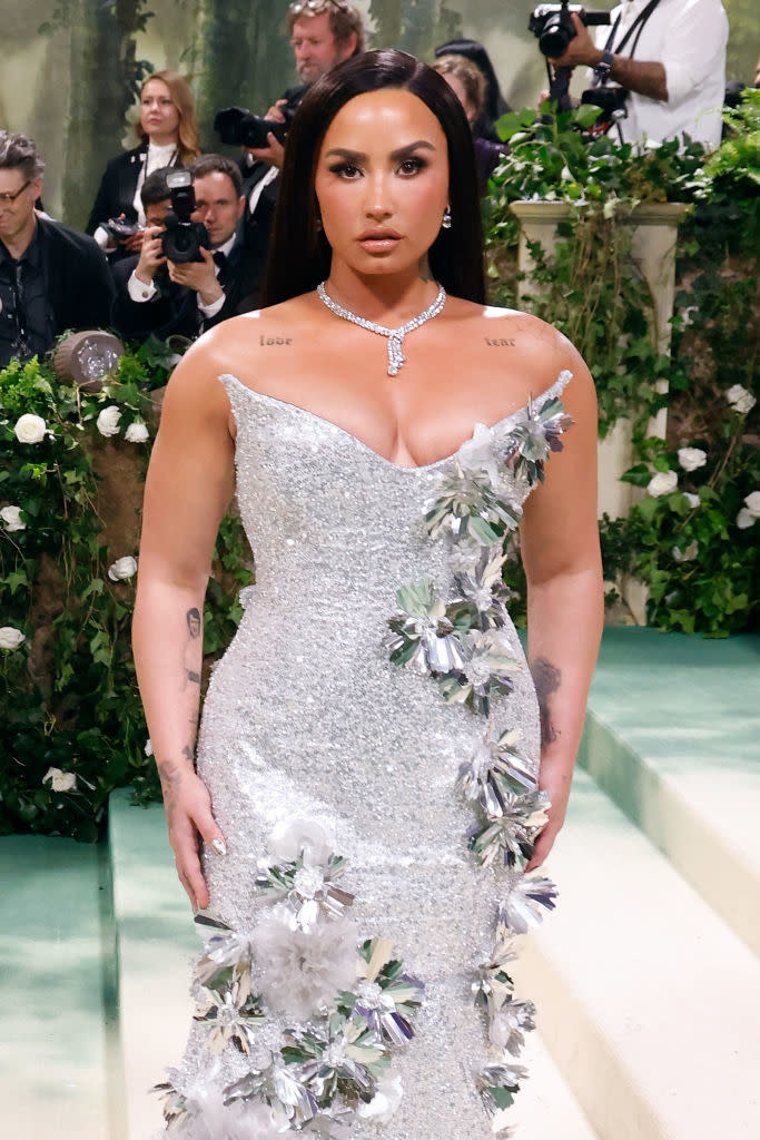 Demi in a sparkling gown with floral embellishments at an event, photographers in background
