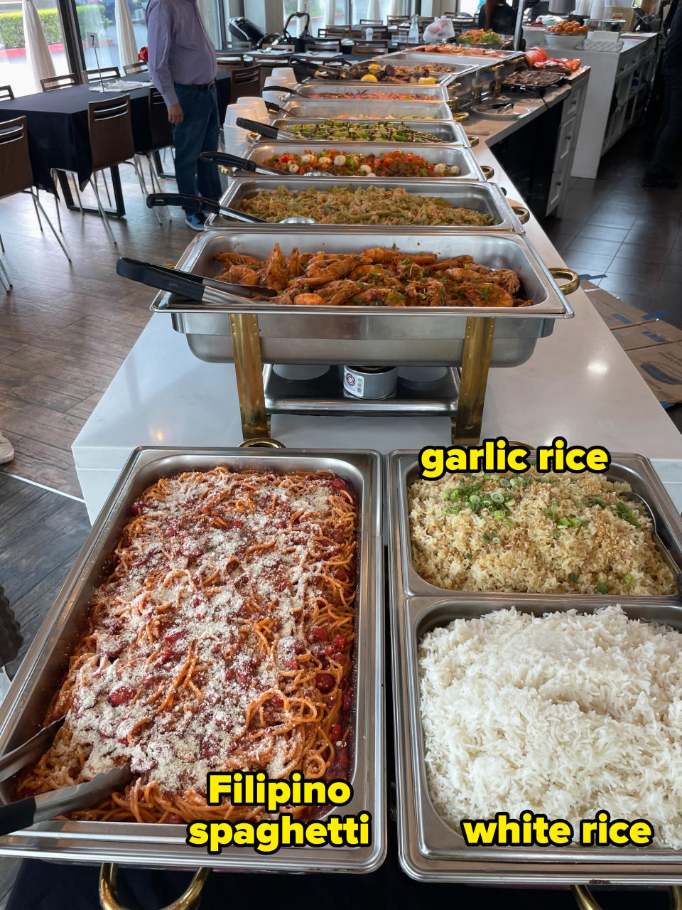 A buffet table with various dishes including spaghetti, rice, fried rice, vegetables, shrimp, and salads. People are serving themselves in the background