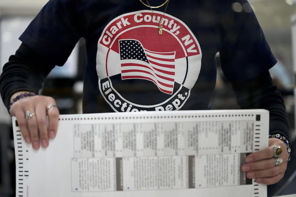 A person wearing a shirt with an American flag and text saying "Clark County Nevada Election Department" holds a sheaf of ballots.