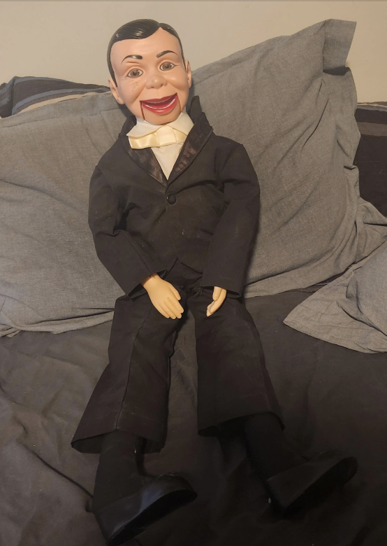 Ventriloquist dummy wearing a suit and bow tie, sitting on a bed propped up against pillows