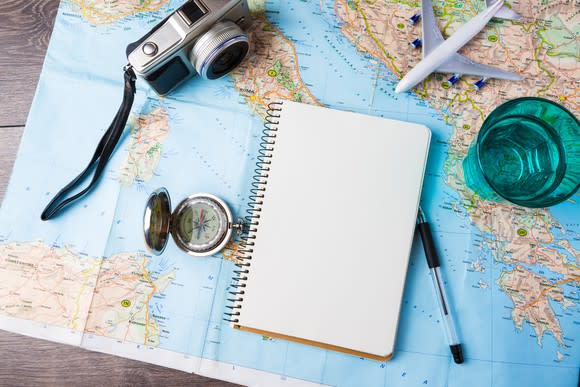 Travel tools on a table including a camera, compas, map, notebook and pen, and a small airplane