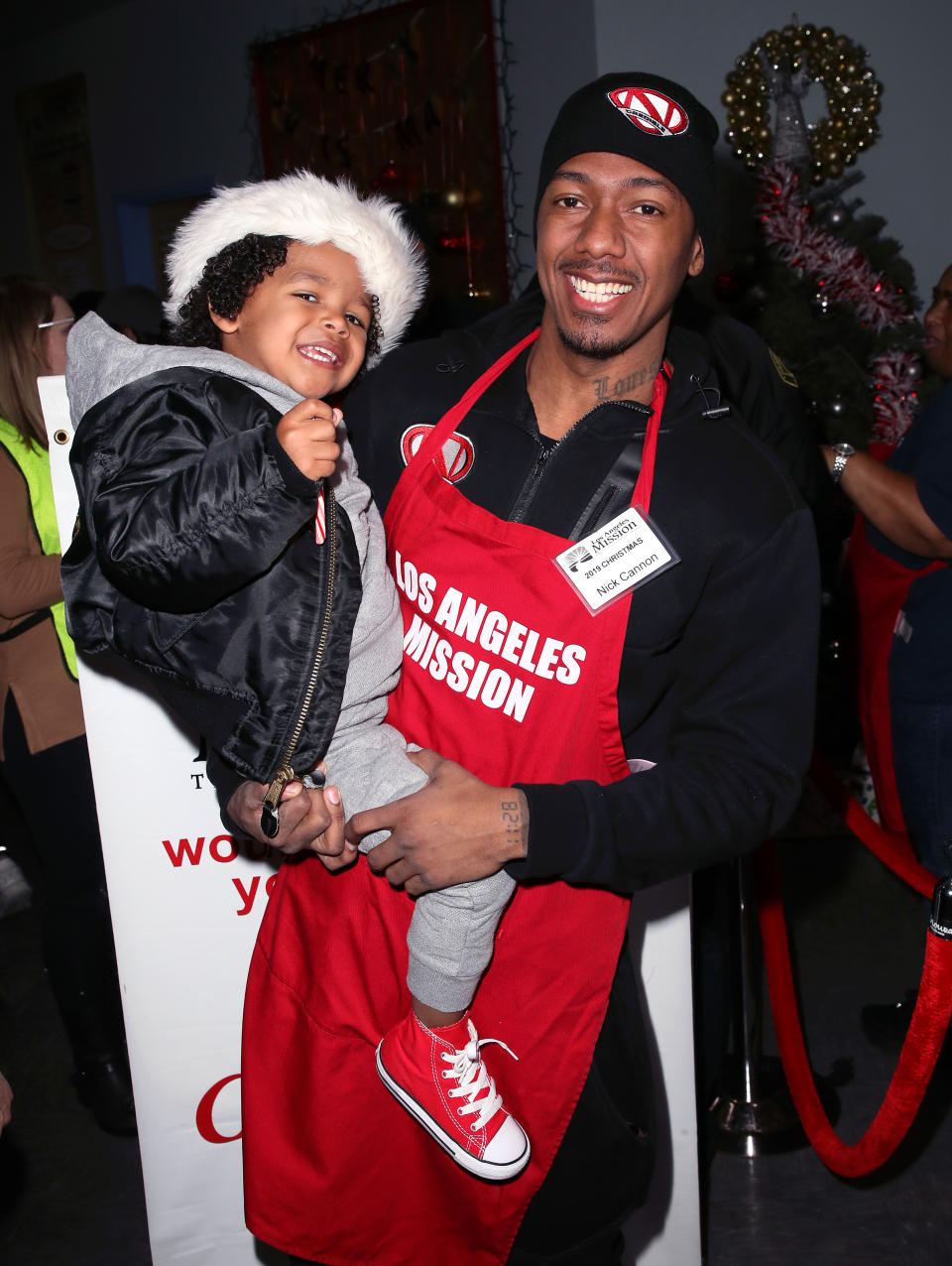 Nick Cannon holds a smiling child while wearing a red "Los Angeles Mission" apron at a charity event