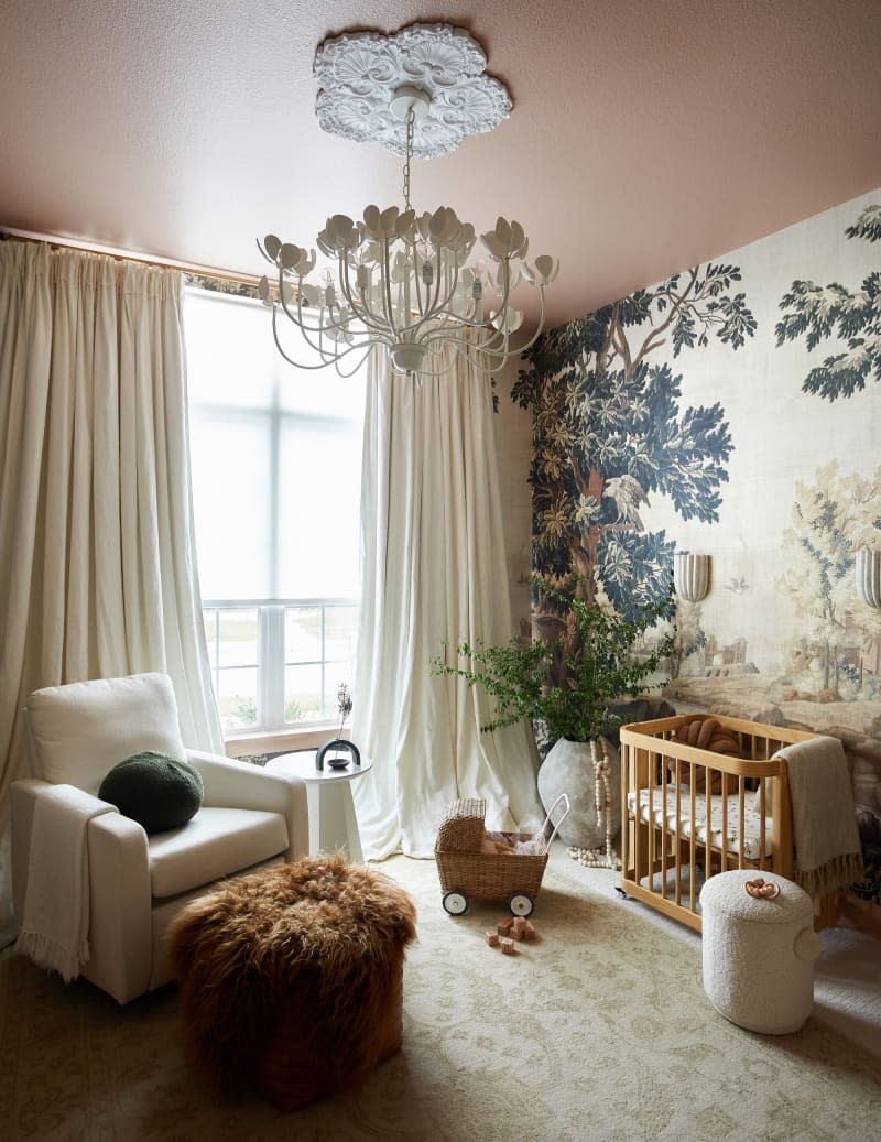 landscape wallpaper accent wall, pink ceiling, floral chandelier with detailed ceiling plate, wood crib, white arm chair, cream curtains