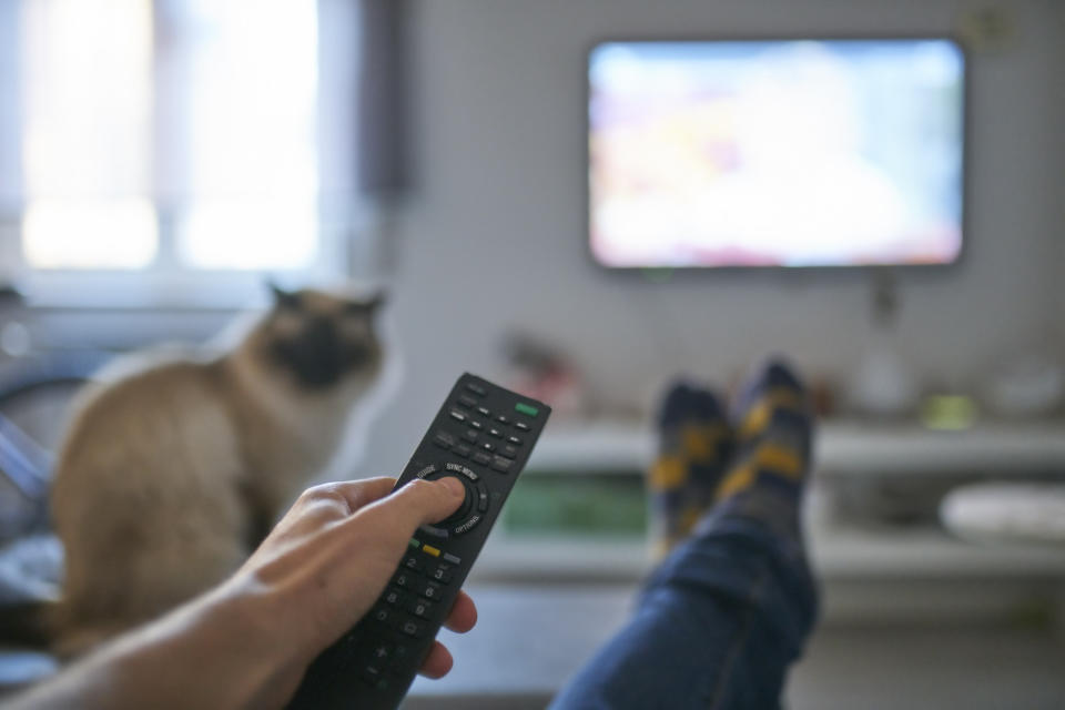 A person's feet up on a table and their hand clicking the remote to a TV, with a cat in the background