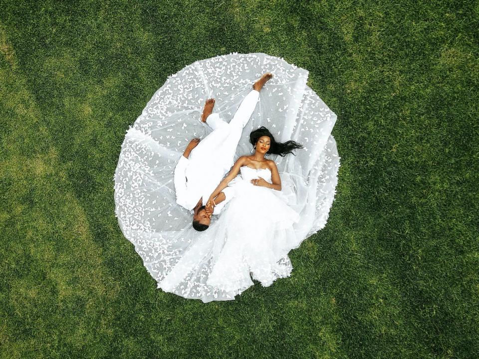 A bride and groom lay on the grass, with her wedding dress spreading around them.