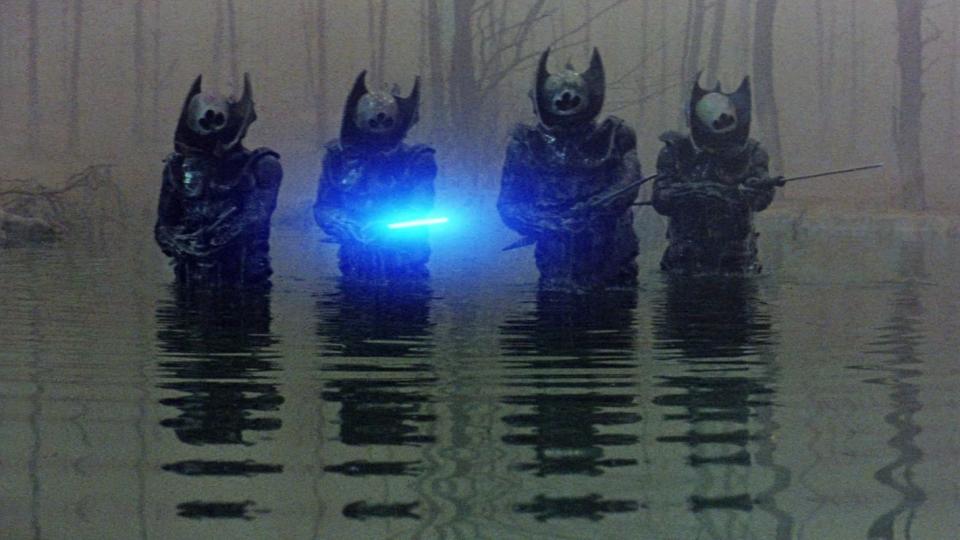 Still from the science fantasy movie Krull (1983). Four evil-looking alien soldiers known as Slayers. They're all wearing spiky black armor and horned helmets, whilst holding sword-like weapons. One soldier is holding a blade that is glowing with bright blue light.