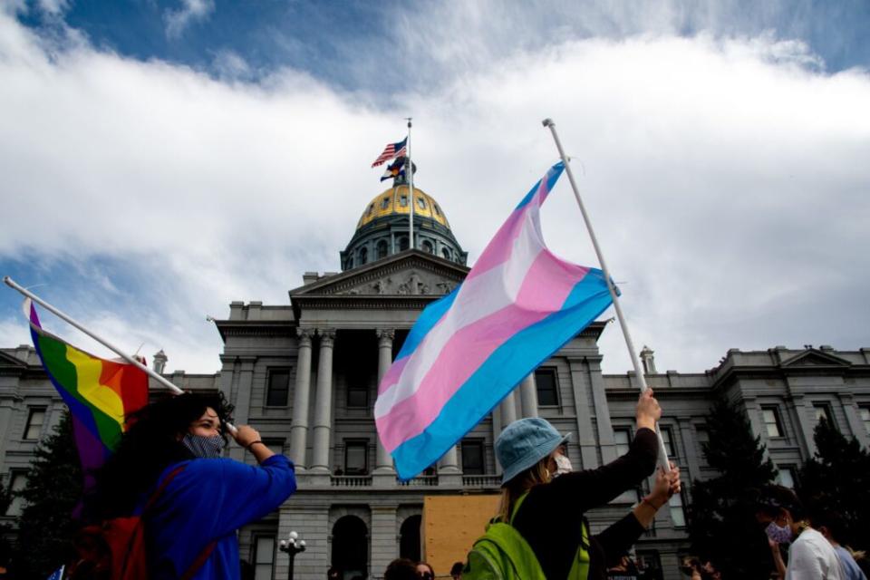 Two people fly a rainbow LGBTQ pride flag and a transgender pride flag in front of the Colorado Capitol building during a celebration