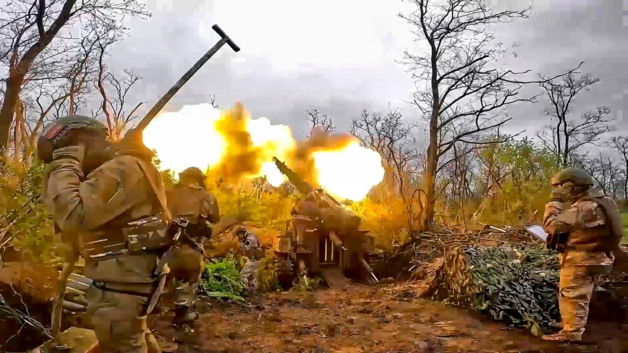 Troops cover their ears as a howitzer fires in a wooded area.