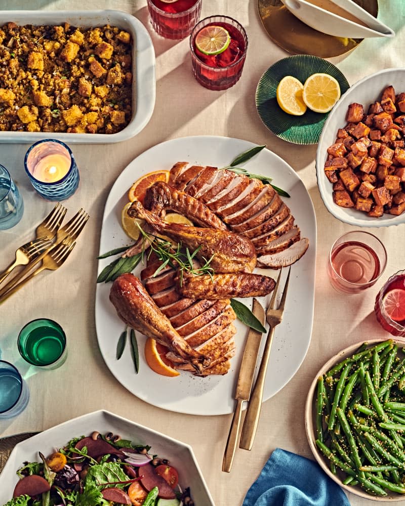 Thanksgiving table with multiple dishes, one with cornbread stuffing, a platter with sliced turkey, a bowl of candied yams, bowl of green beans. Forks and glasses on table