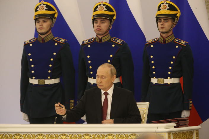 President Vladimir Putin prepares to sign a document as three guards in formal uniforms with brass buttons and peaked caps stand to attention behind him.