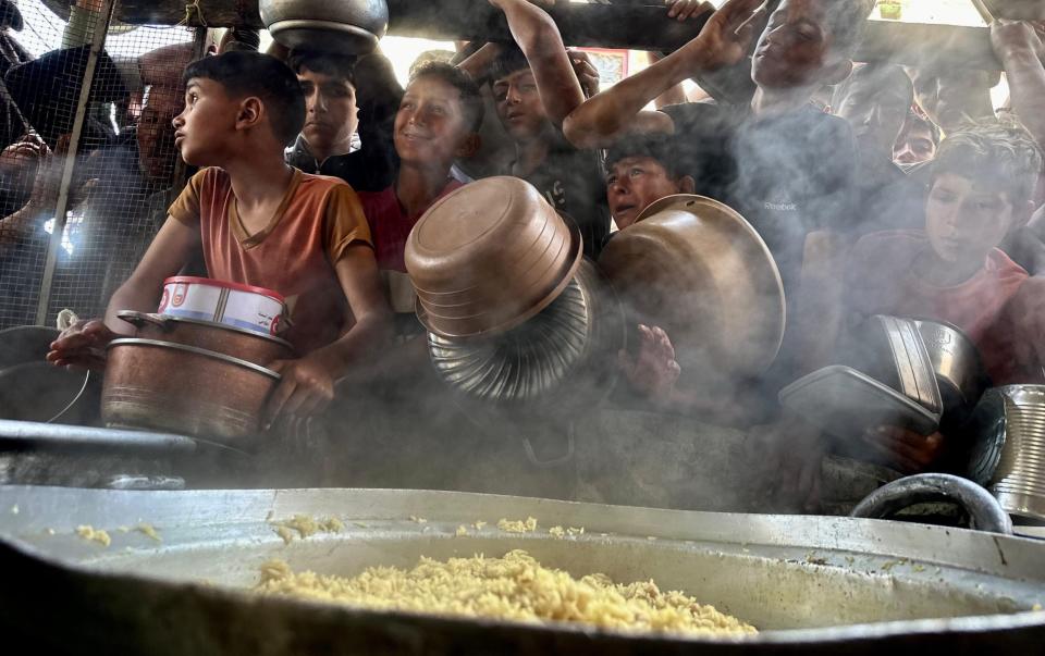 Children holding bowls queue for food in front of a steaming pan of rice