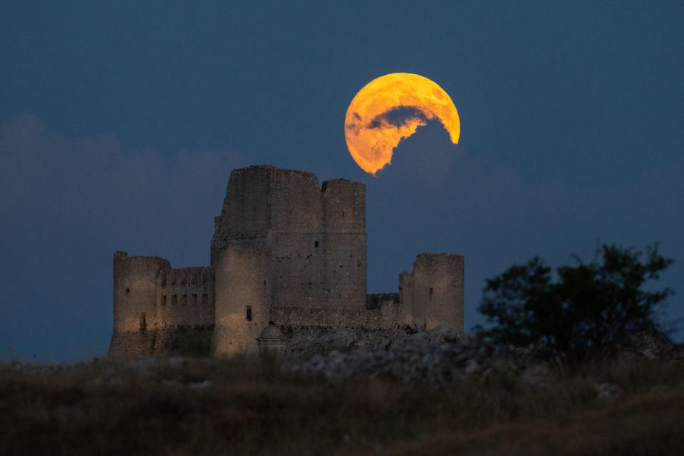 bright golden full moon partially obscured by clouds above a ruin of a large castle.