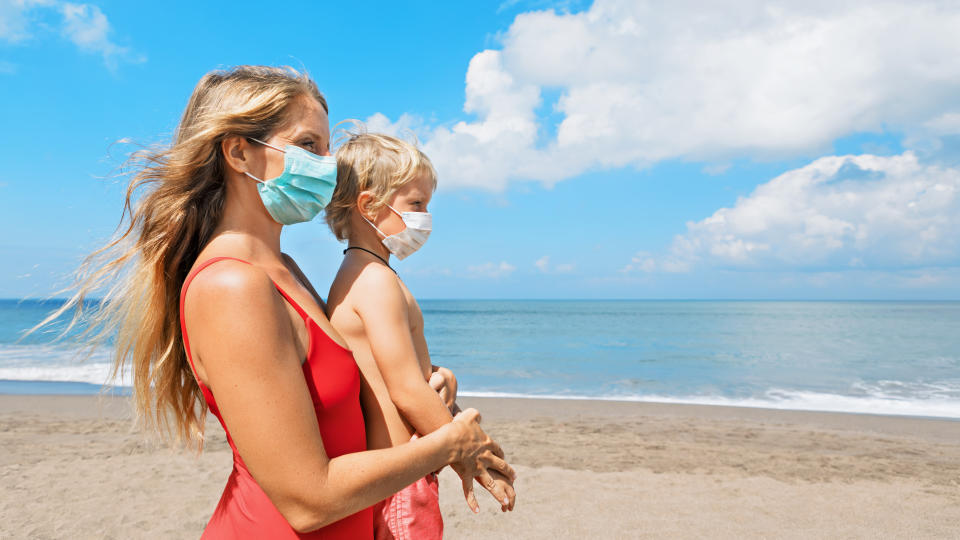 Mother put medical mask on child on sea beach. New rules to wear cloth face covering at public places. Cancelled cruise, tour due coronavirus COVID 19. Family vacation, travel lifestyle at summer 2020