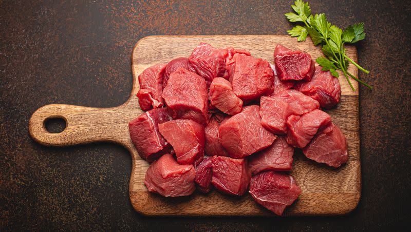 Raw beef meat chopped in cubes with fresh parsley sits on a wooden cutting board for cooking stew or other meat dish. How much red meat one consumes impacts some health factors.