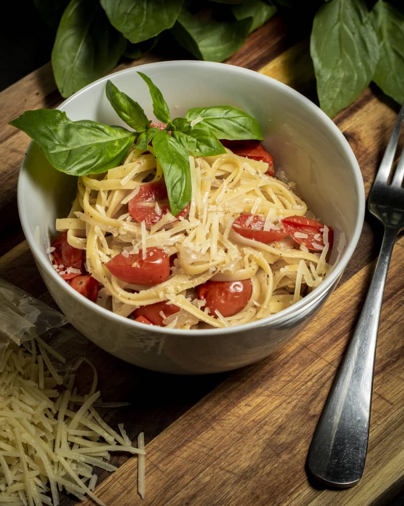 bowl of spaghetti with cherry tomatoes and basil leaves on a wooden table with ingredients positioned in frame