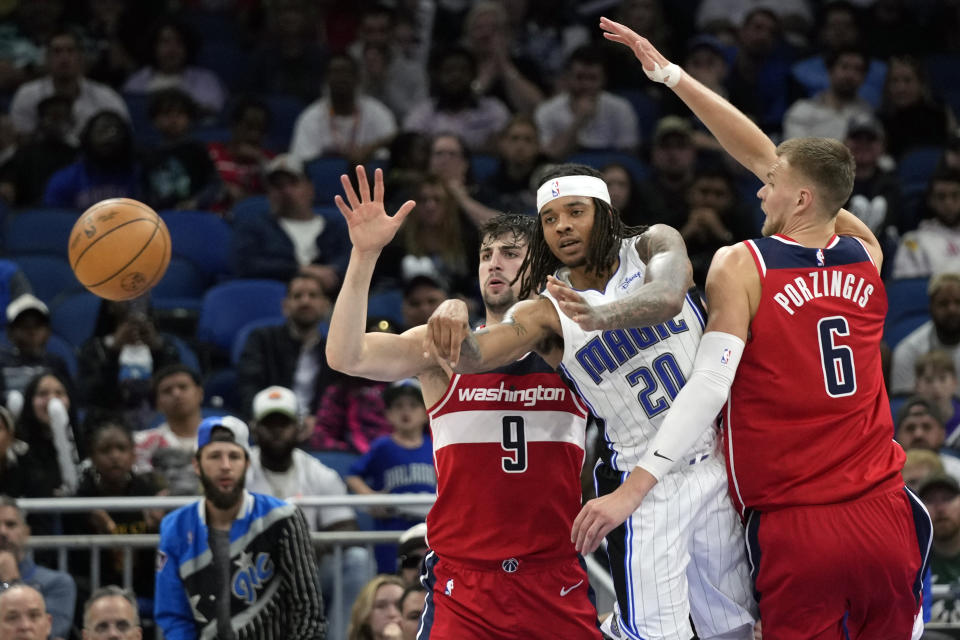 Orlando Magic's Markelle Fultz (20) passes the ball as gets caught between Washington Wizards' Deni Avdija (9) and Kristaps Porzingis (6) during the second half of an NBA basketball game, Tuesday, March 21, 2023, in Orlando, Fla. (AP Photo/John Raoux)