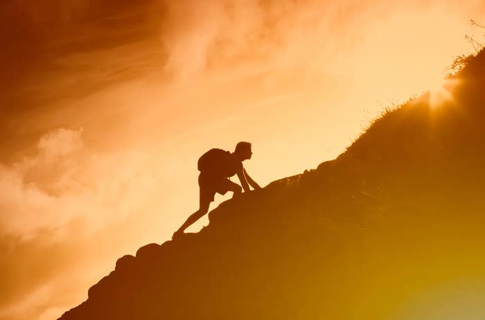 A person with a backpack is climbing a steep, rocky hill against a dramatic sky, silhouetted by the sunlight