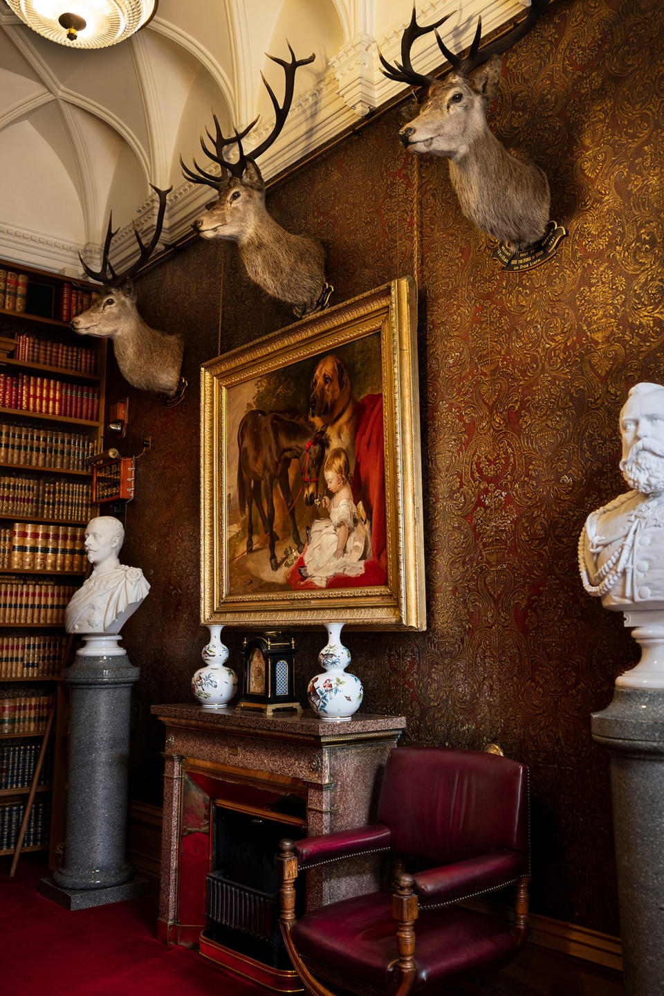 A room in Balmoral featuring book shelves, a painting over a fireplace and three stag heads on a wall