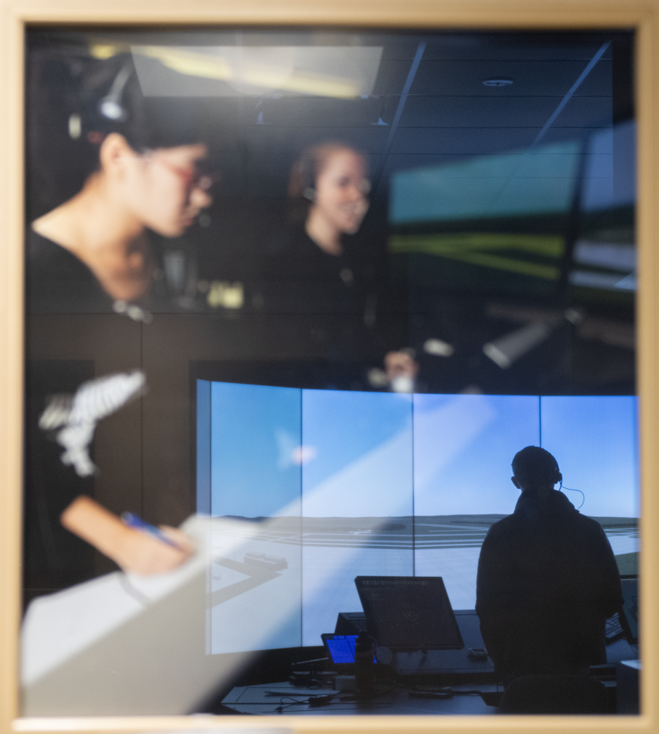 Looking through the window of the Aeronautics Traffic Control Simulator as a  student works on the tower simulator in the lower level called the Bot Bunker.